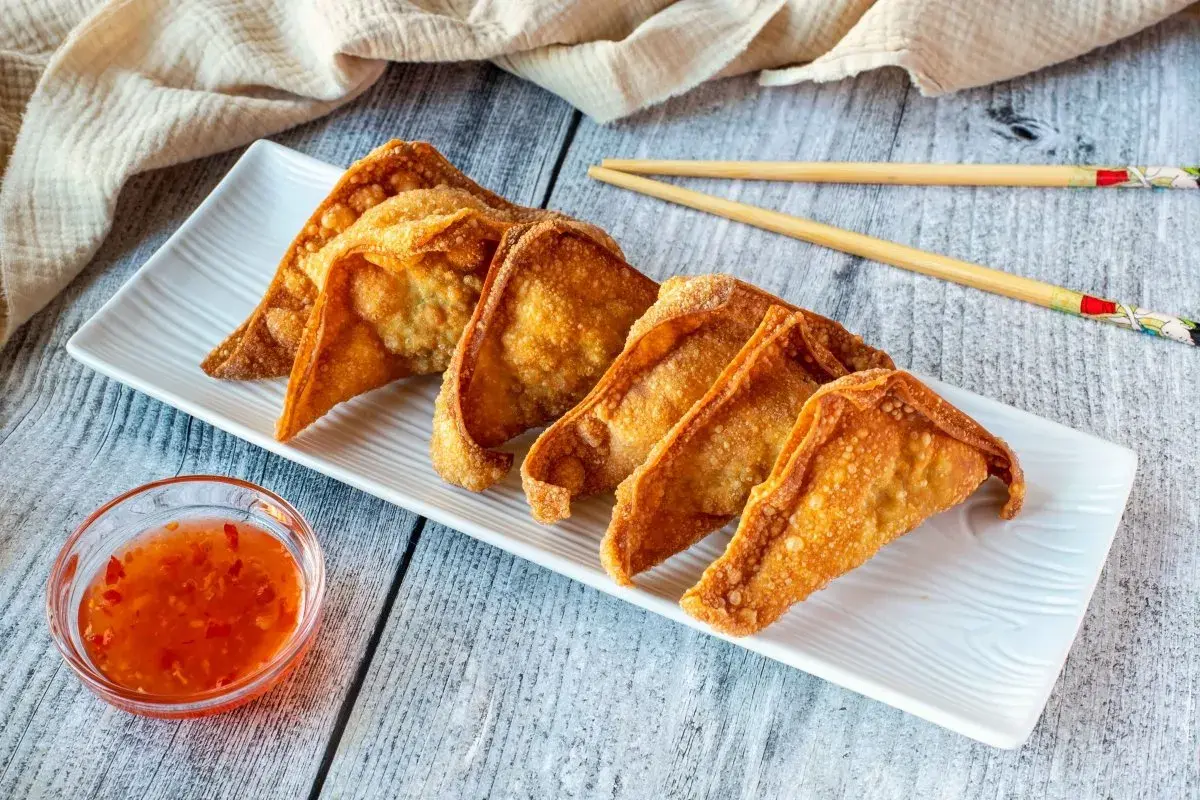 Crujientes wan tun frito dorados en una bandeja blanca, acompañados de salsa agridulce y palillos.