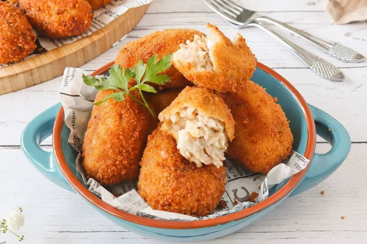 Croquetas de atún doradas y crujientes, servidas en un cuenco azul. Una está partida, mostrando su cremoso interior.