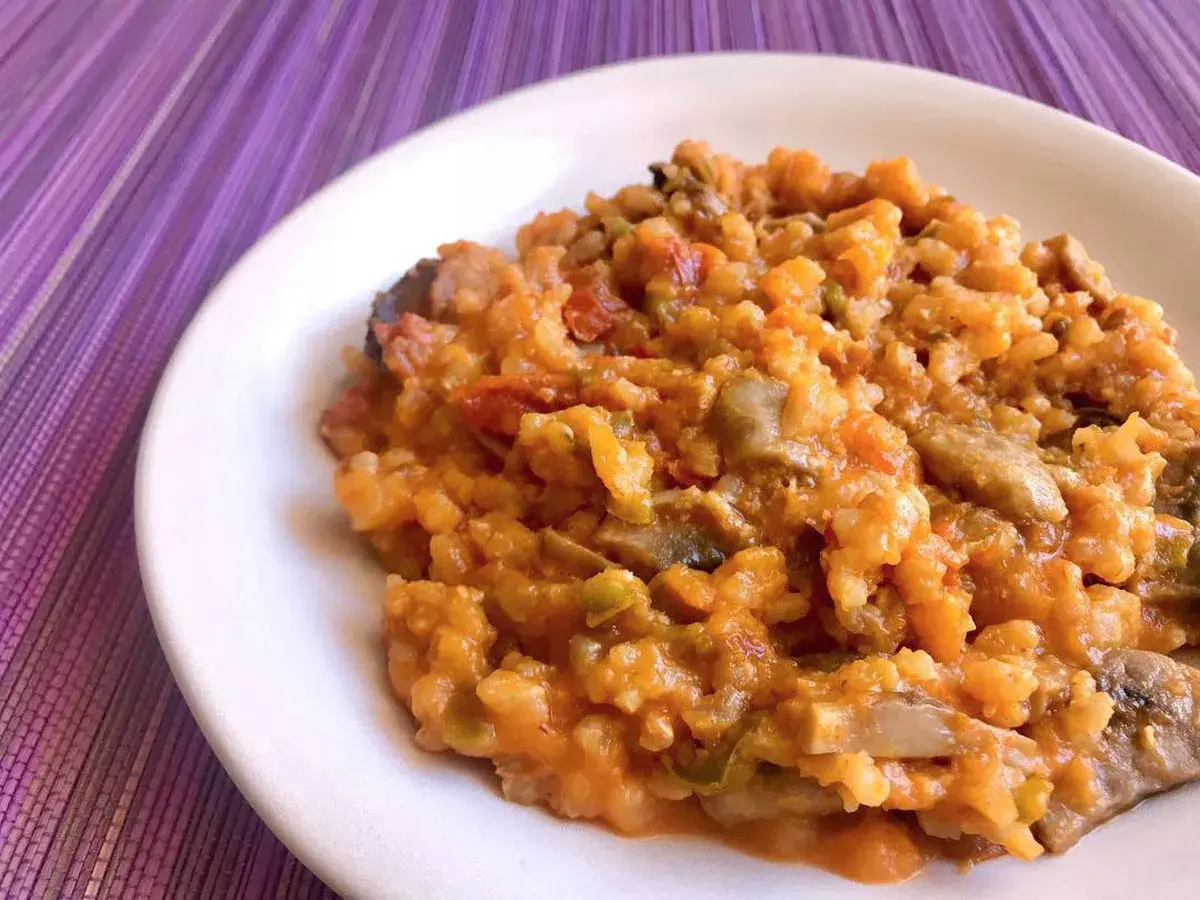 Un cremoso risotto de calabaza con trozos de champiñones y pimientos, servido en un plato blanco sobre un mantel morado.