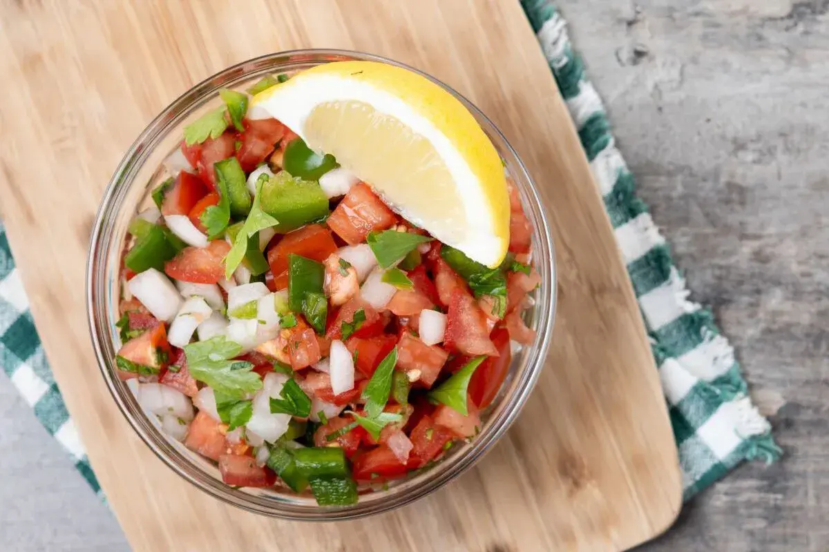 Un tazón de pico de gallo fresco, con tomate, cebolla, pimiento verde y cilantro, adornado con una rodaja de limón.