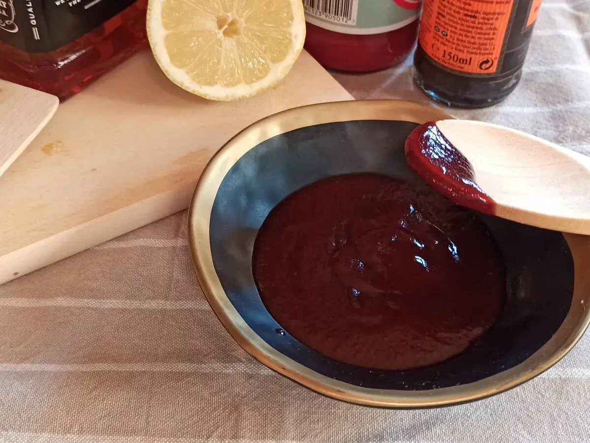 Cuchara de madera prueba la deliciosa salsa barbacoa casera en un cuenco. Limón y botellas de ingredientes al fondo.