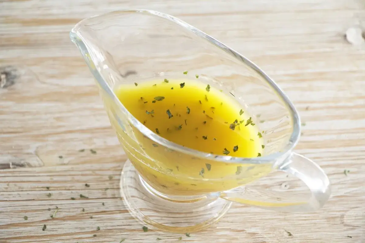 Vinagreta de mostaza dorada con hierbas verdes picadas, servida en una salsera de cristal sobre una mesa de madera.