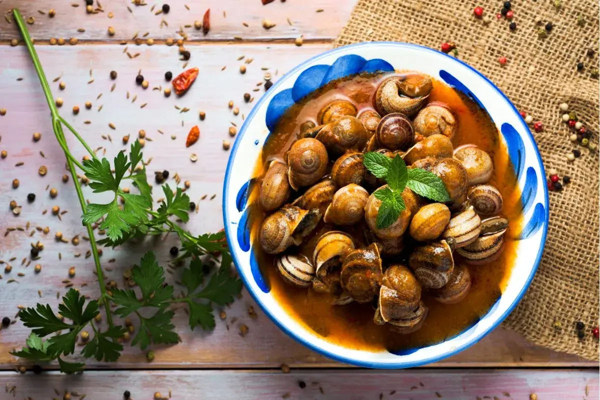 Un plato de caracoles en salsa, adornado con menta fresca, rodeado de especias y perejil sobre una mesa de madera.