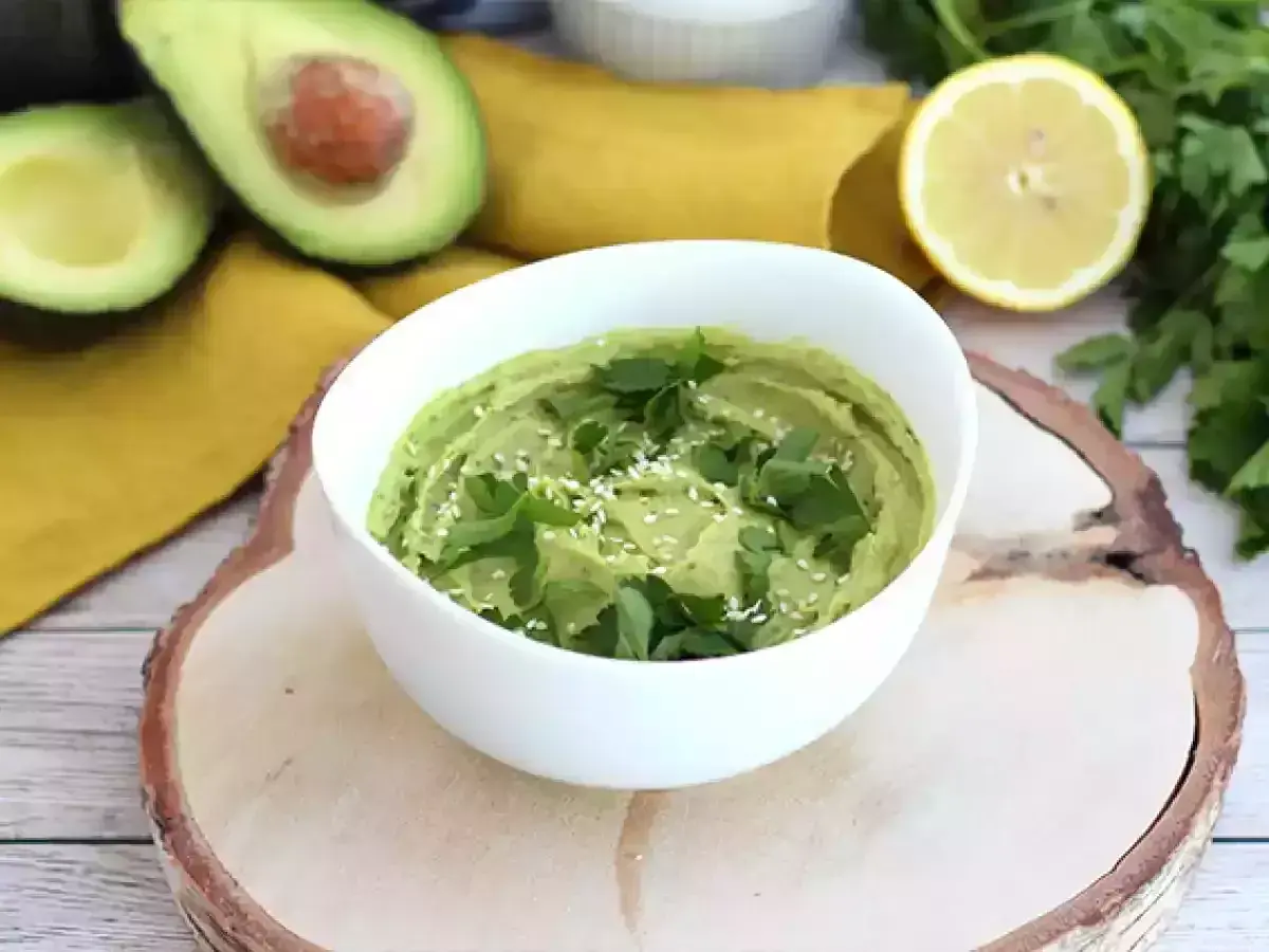 Un cremoso hummus de aguacate, decorado con perejil fresco y semillas de sésamo, junto a aguacates cortados y un limón.