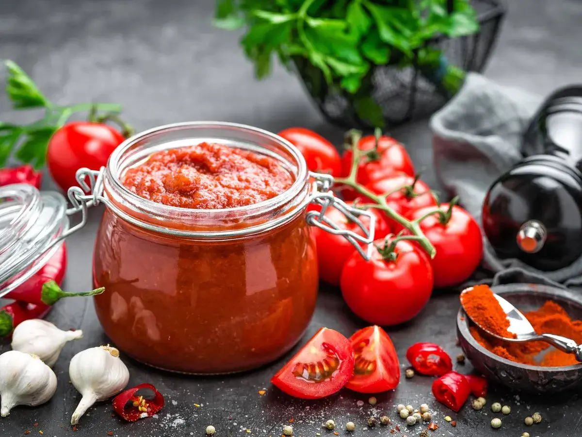 Frasco de vidrio lleno de vibrante salsa de tomate casera, rodeado de tomates frescos, ajos, chiles y especias.