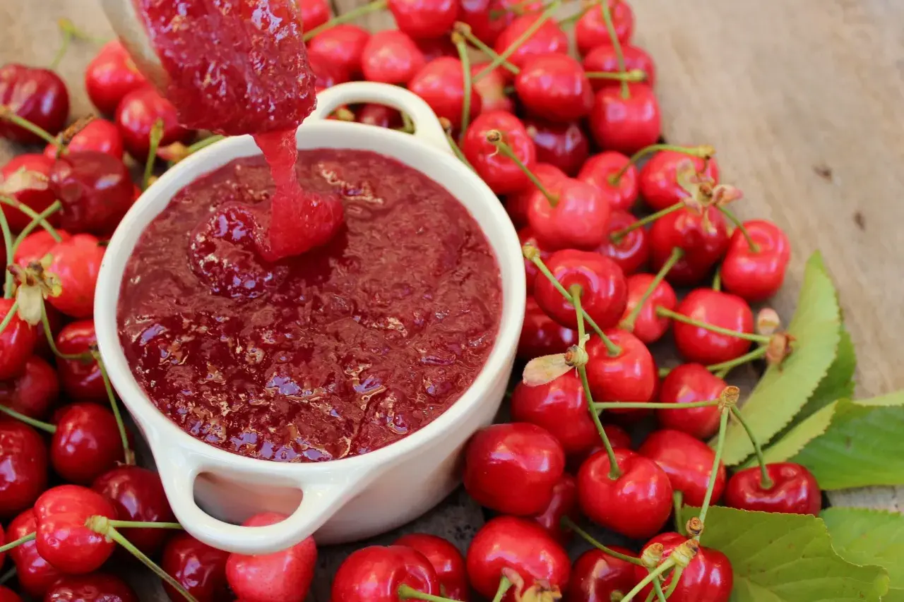 Cuchara vierte mermelada de cerezas casera en un cuenco blanco, rodeado de cerezas frescas y hojas verdes.
