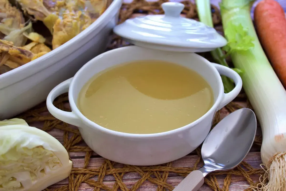 Un tazón humeante de caldo de pollo casero, listo para reconfortar. Verduras frescas como puerro, zanahoria y repollo acompañan.