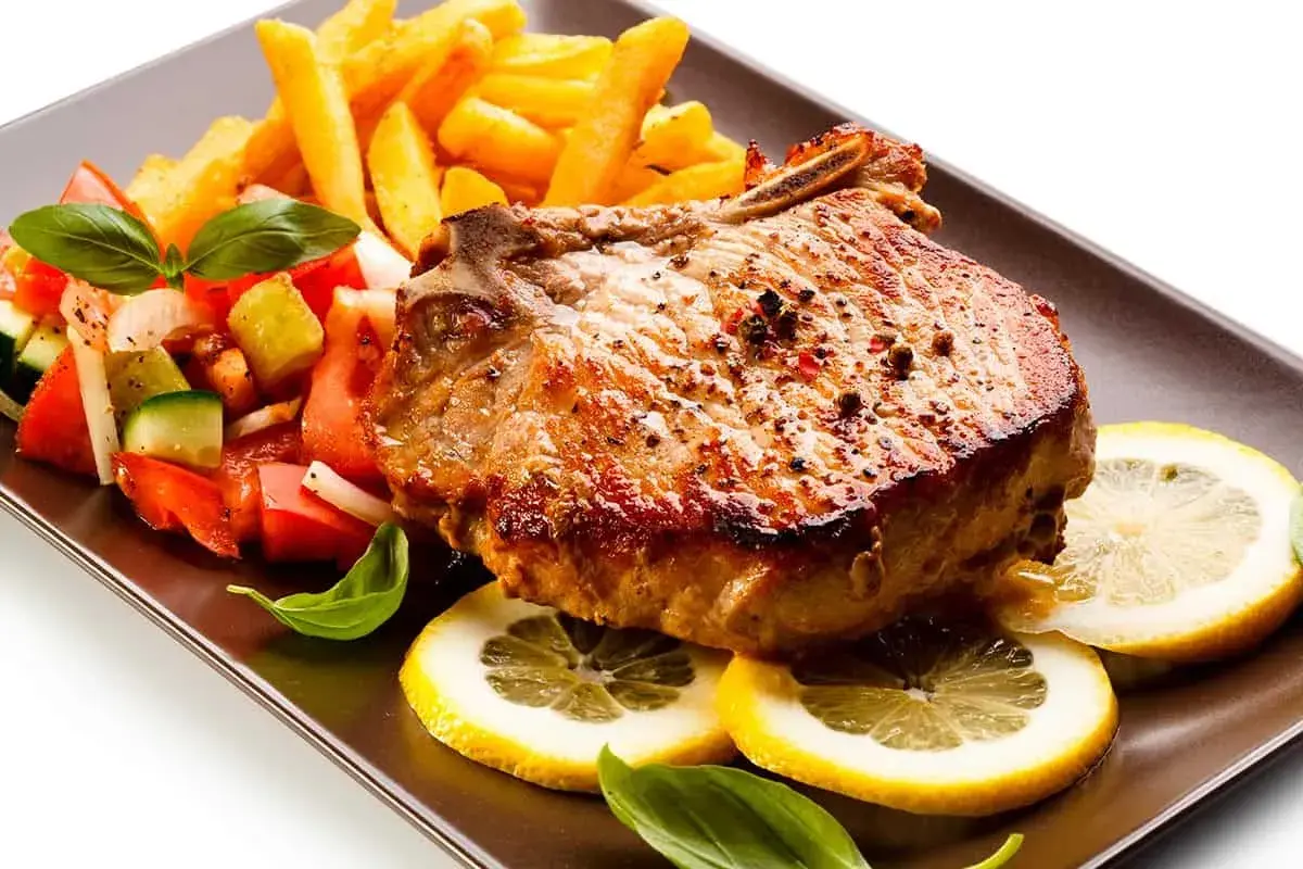 Chuletas de cerdo al horno jugosas, acompañadas de papas fritas crujientes, ensalada fresca y rodajas de limón.