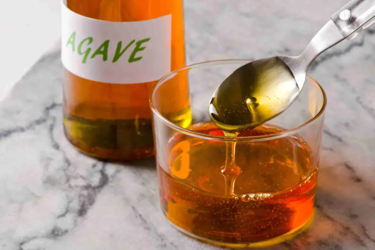 Cuchara con sirope de agave cayendo en un vaso, junto a una botella etiquetada.