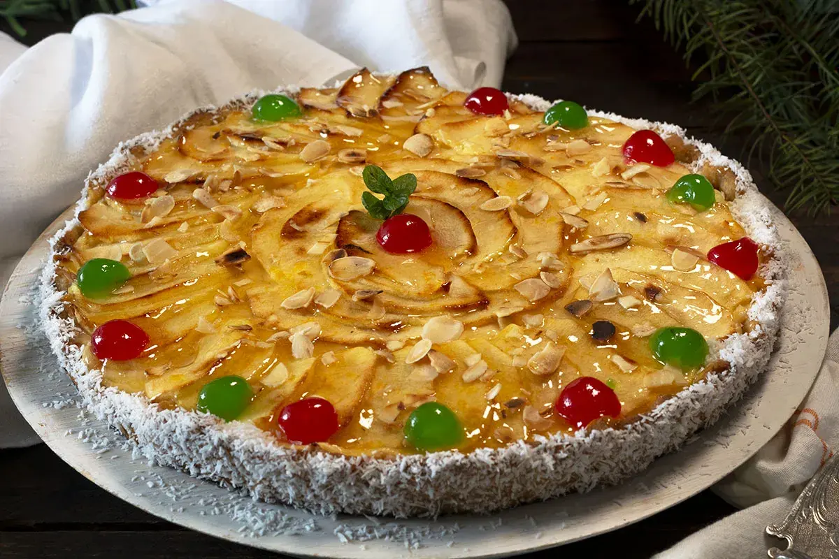 Tarta de manzana con crema pastelera, decorada con almendras laminadas, cerezas rojas y verdes, y coco rallado en el borde.