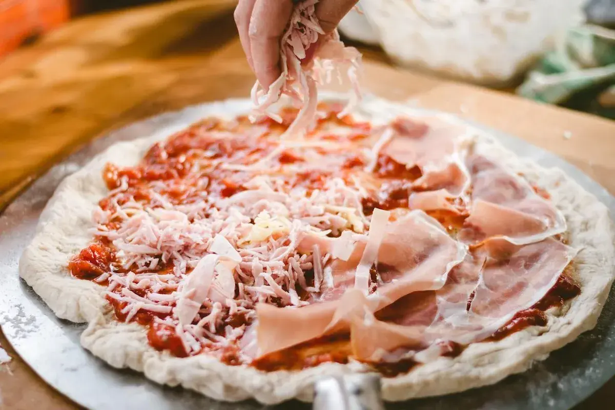 Mano esparciendo queso rallado sobre una masa de pizza casera con jamón y salsa de tomate.