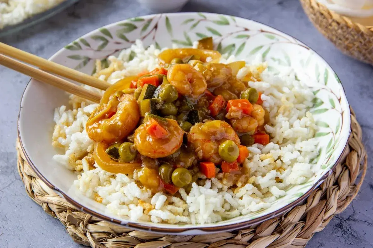 Un plato de arroz blanco cubierto con gambas en salsa agridulce y verduras. Los palillos descansan sobre el borde del cuenco.