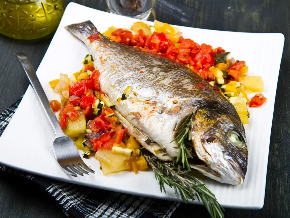 Dorada al horno con verduras asadas, servida en plato blanco con tenedor y romero fresco.