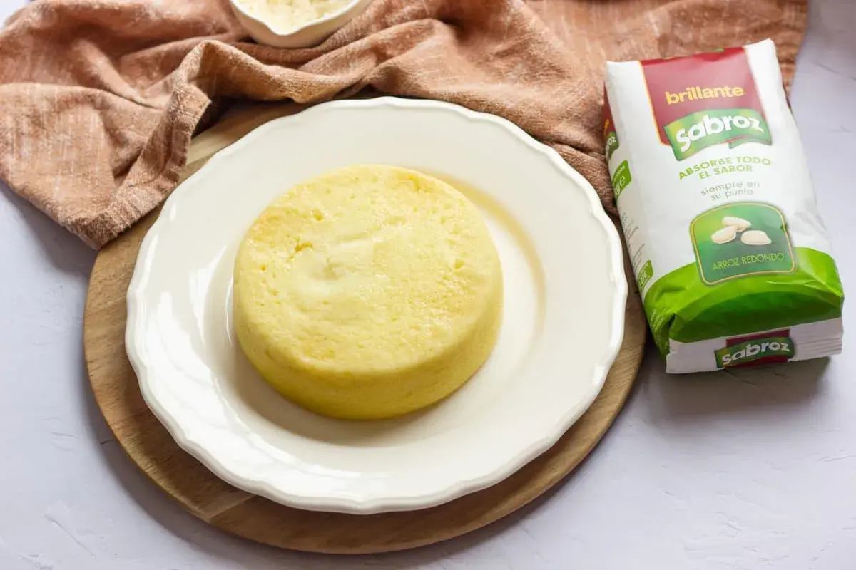 Pastel de arroz amarillo, esponjoso y redondo, servido en plato blanco junto a un paquete de arroz Sabroz.
