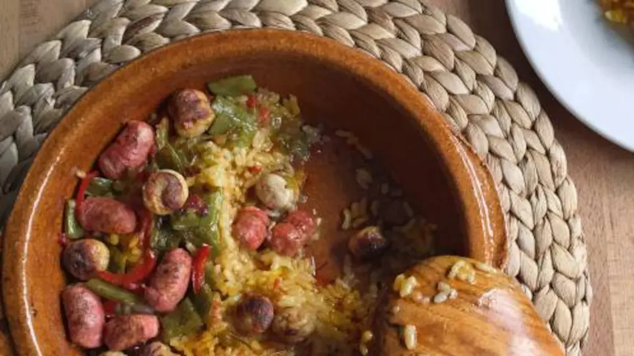 Un plato de barro con arroz, salchichas, pimientos verdes y rojos. Un cucharón de madera reposa en el borde.