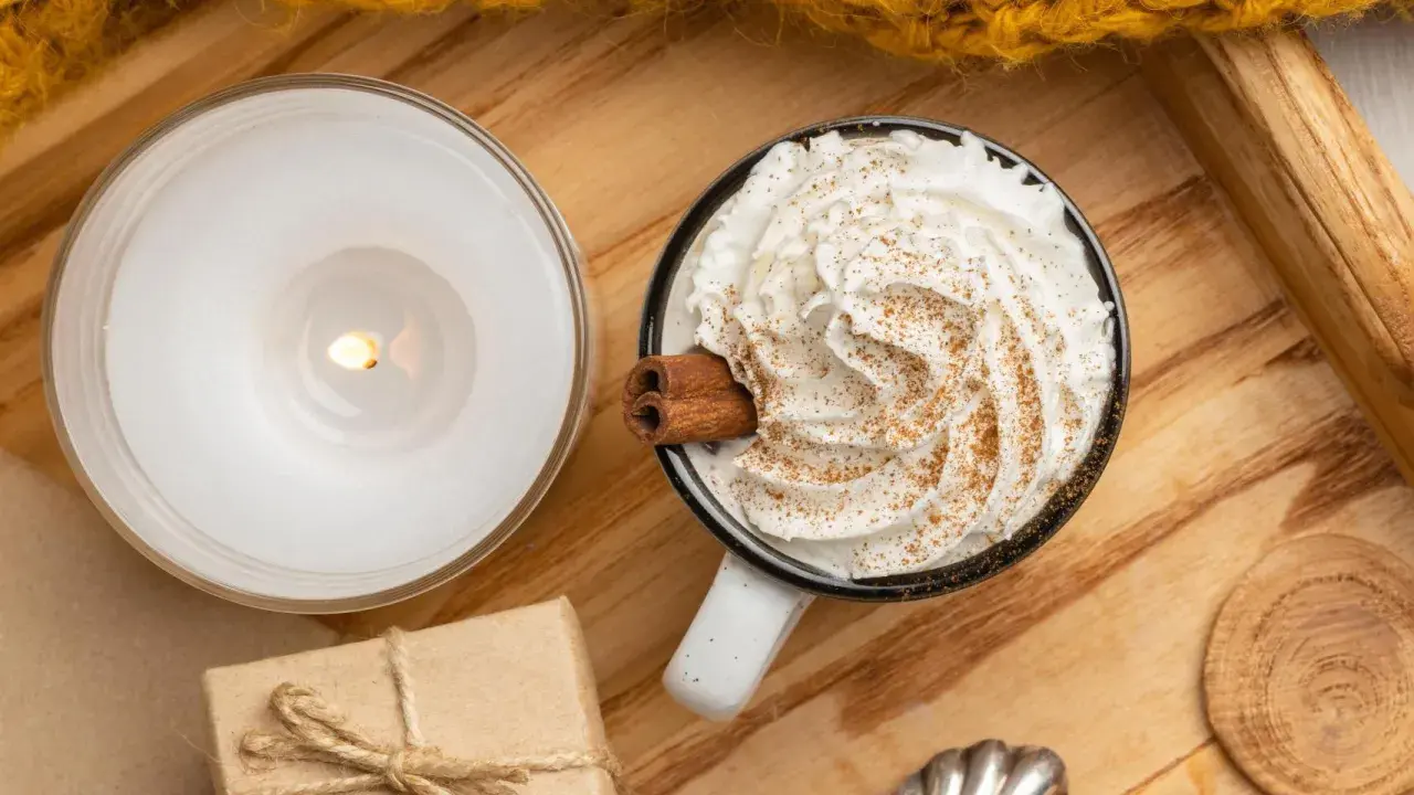 Taza de chocolate caliente con abundante crema de leche, canela y una rama de canela. Una vela encendida y un regalo completan la escena acogedora.