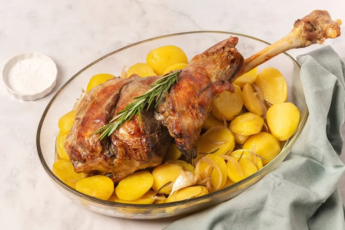 Pierna de cordero al horno dorada y jugosa, acompañada de patatas asadas y romero en una fuente de cristal.