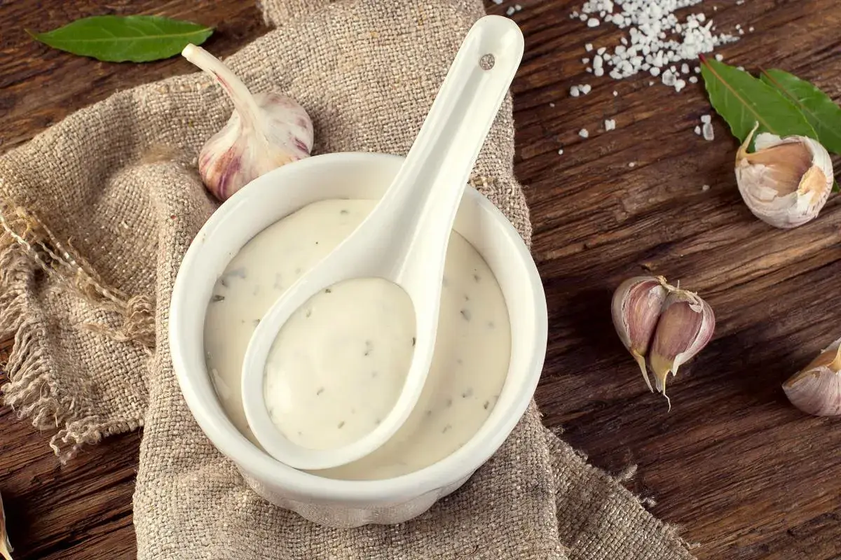Crema de salsa de ajo en un cuenco blanco con una cuchara, rodeada de ajos y hojas verdes sobre una mesa de madera rústica.