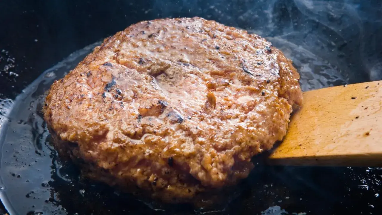 Jugosa hamburguesa de ternera dorándose en sartén, con vapor ascendiendo y una espátula de madera lista para darle la vuelta.