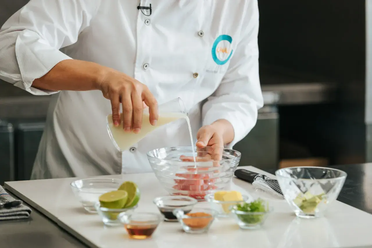 Chef vierte líquido en un bol con pescado, preparando la leche de tigre receta. Ingredientes frescos como limas y aguacate a la vista.