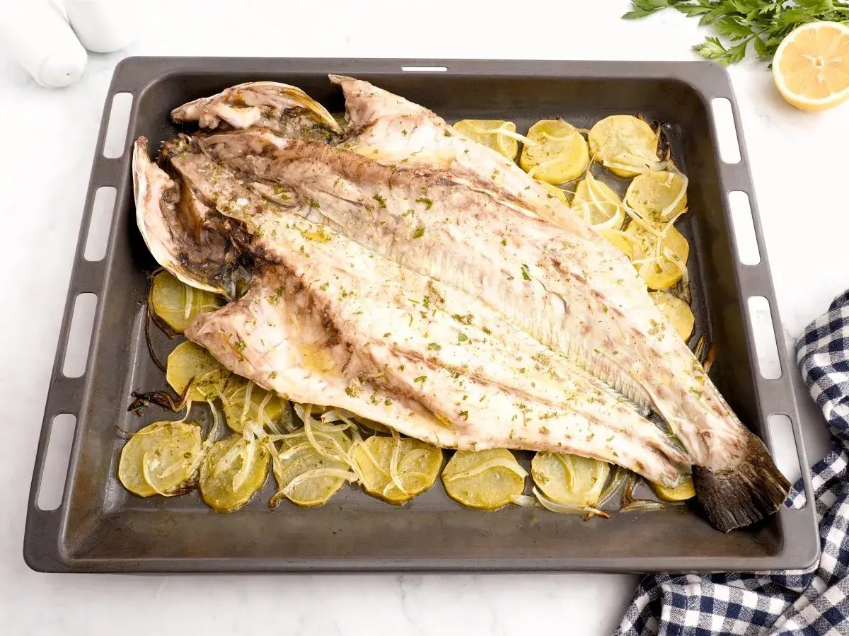 Corvina al horno, entera y dorada, reposa sobre rodajas de patata y cebolla en una bandeja.