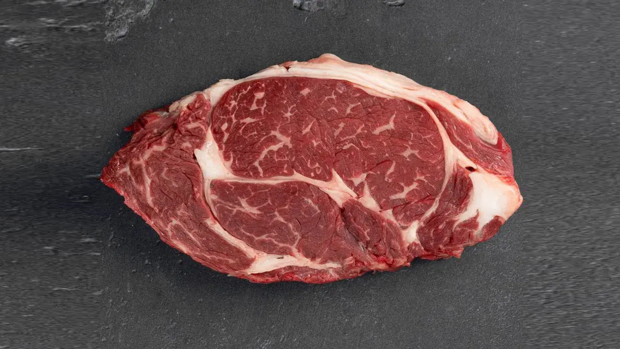 Un corte de aguja de ternera cruda, con marmoleo de grasa blanca entre la carne roja, sobre una superficie oscura.