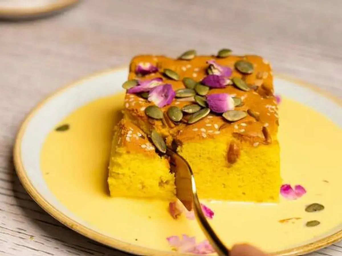 Un trozo de bizcocho de calabaza, decorado con semillas de calabaza y pétalos de rosa, servido en una crema amarilla.