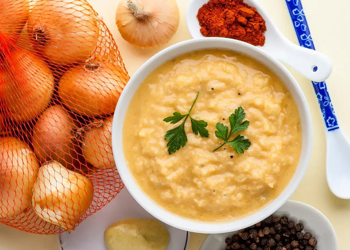 Un tazón de cremosa salsa de cebolla, decorado con perejil, junto a cebollas frescas, pimentón y granos de pimienta.