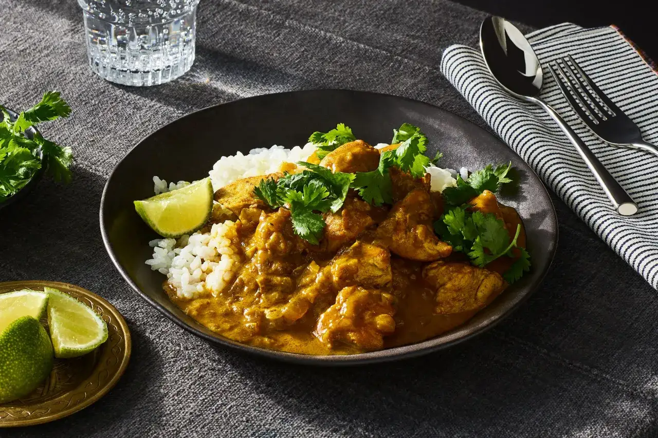 Un delicioso pollo al curry con arroz blanco, cilantro fresco y gajos de lima en un plato oscuro.