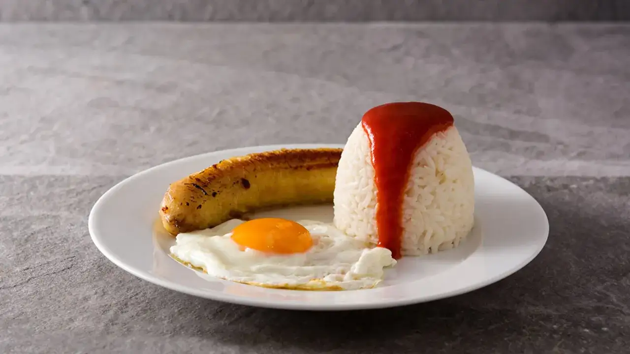 Arroz a la cubana: arroz blanco con salsa roja, huevo frito y plátano maduro frito en un plato blanco.