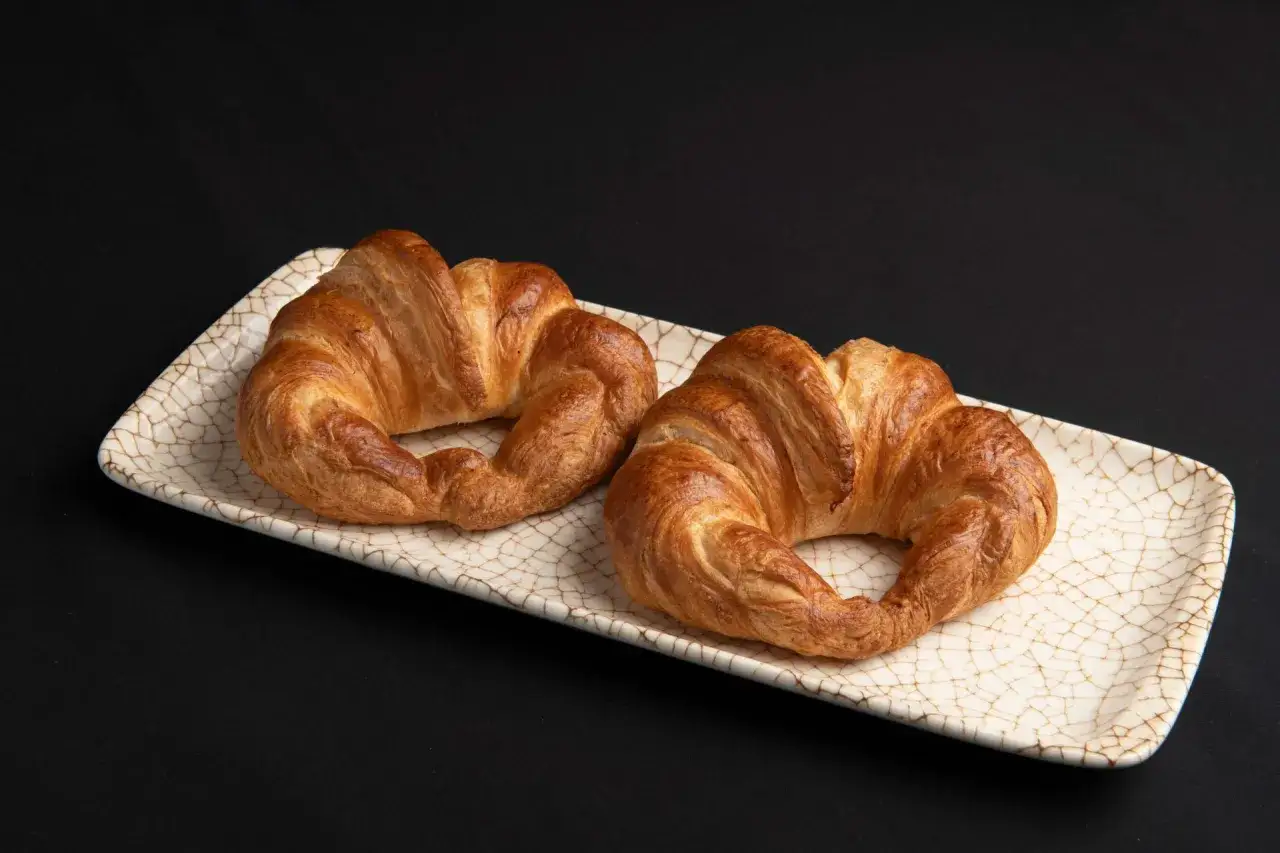 Dos croissants de hojaldre dorados y crujientes reposan en una bandeja de cerámica con un diseño de grietas.