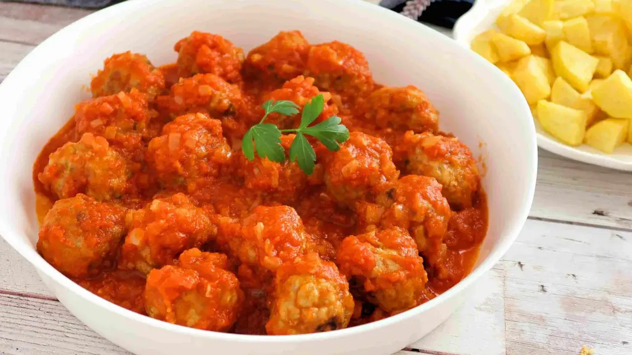 Un tazón blanco lleno de deliciosas albóndigas con tomate, adornado con una ramita de perejil. Al fondo, un plato de patatas fritas doradas.