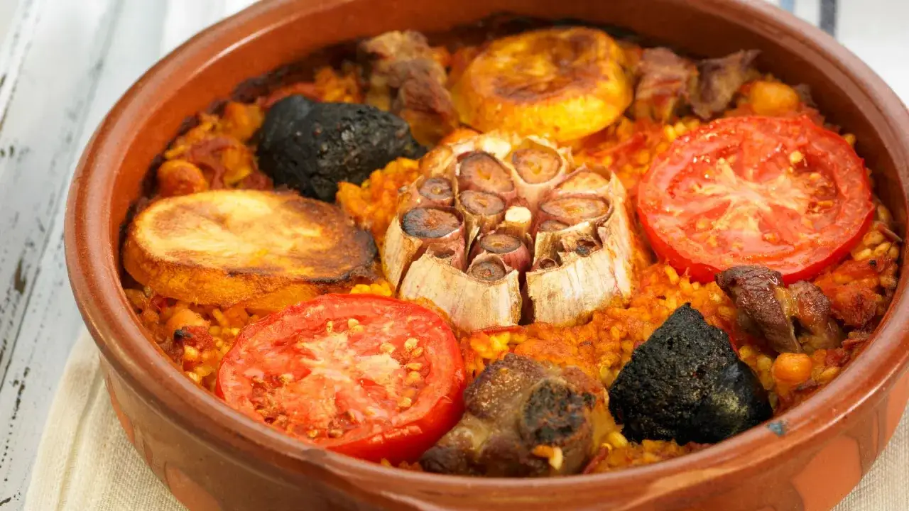 Un sabroso plato de garbanzos con arroz, adornado con rodajas de tomate, patatas fritas, ajo asado y trozos de carne.