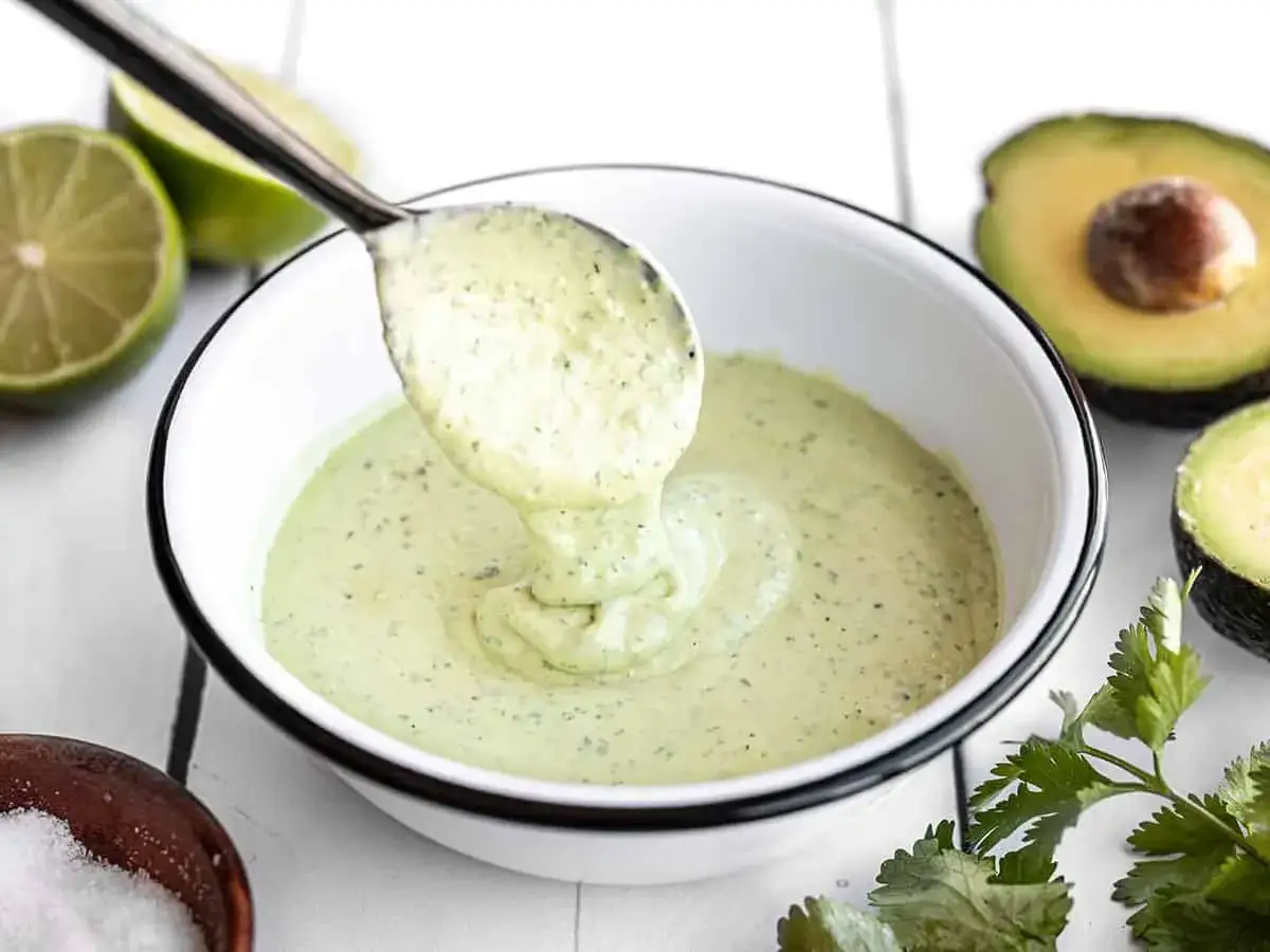Cremosa mayonesa de aguacate servida en un tazón blanco, con limones y cilantro fresco alrededor.