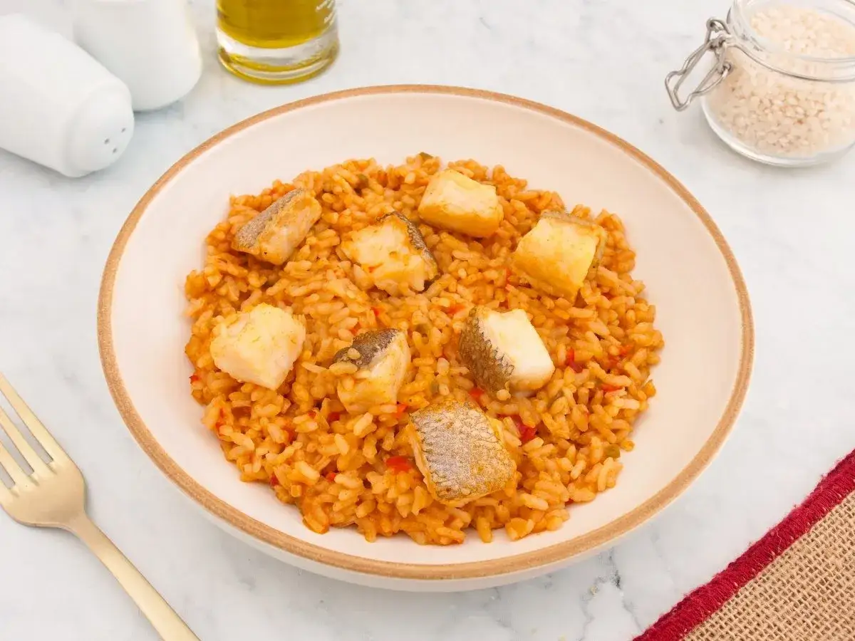 Un plato de arroz con bacalao, con trozos de pescado blanco sobre arroz naranja, adornado con pimientos rojos.