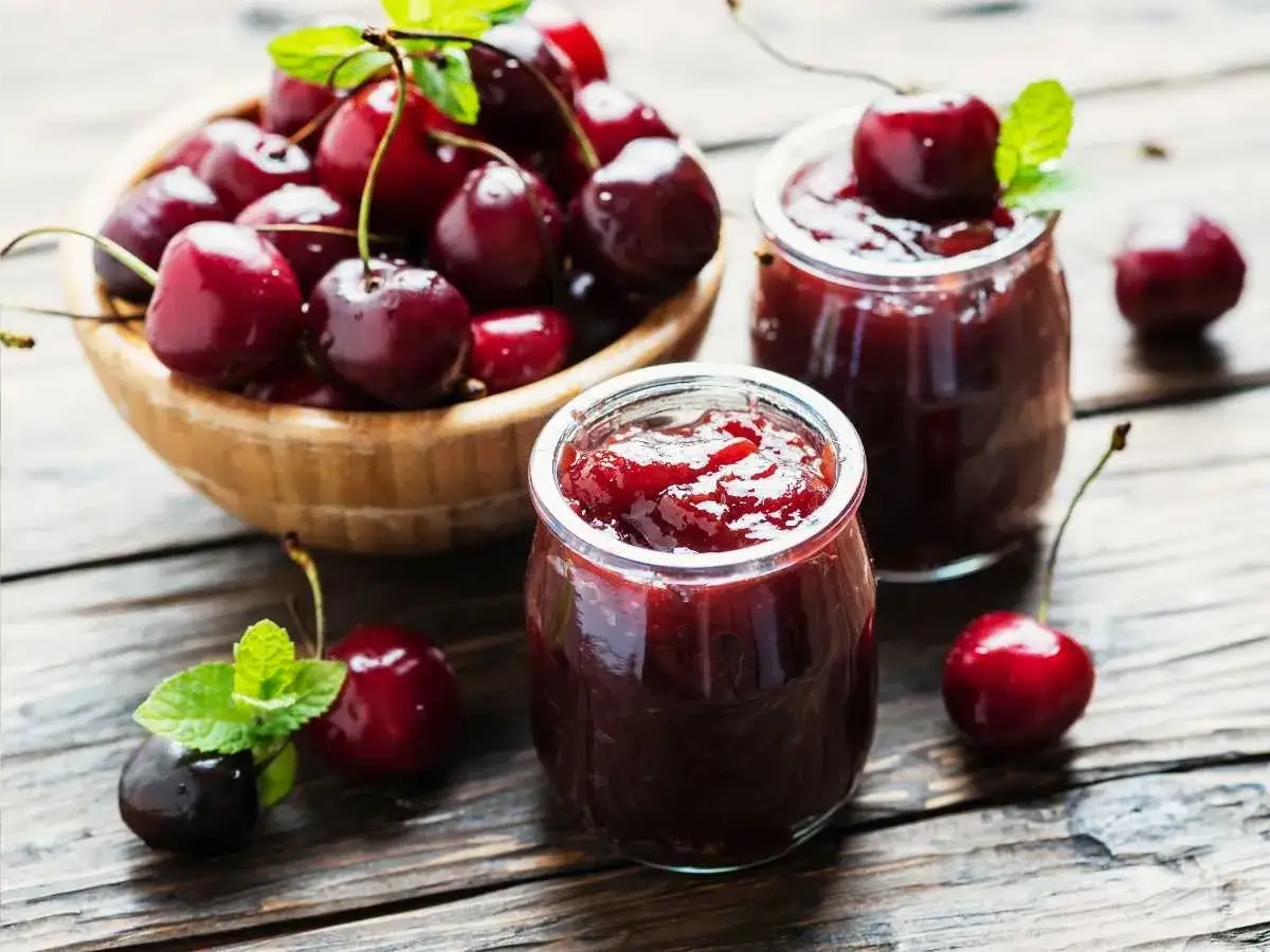 Dos frascos de mermelada de cerezas casera, con cerezas frescas y hojas de menta sobre una mesa de madera rústica.
