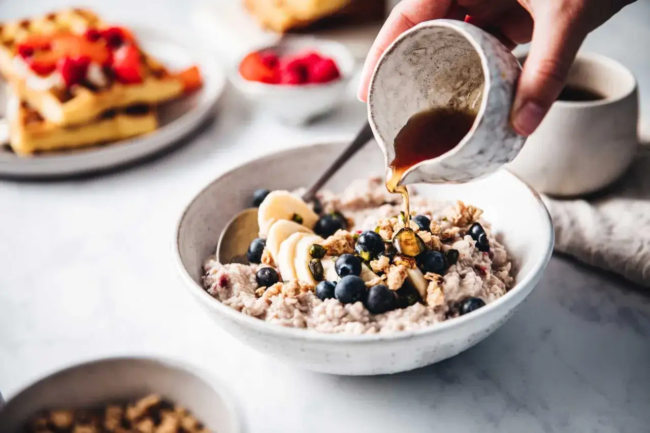A mano vierte sirope de agave sobre un tazón de avena con arándanos, plátano y granola.