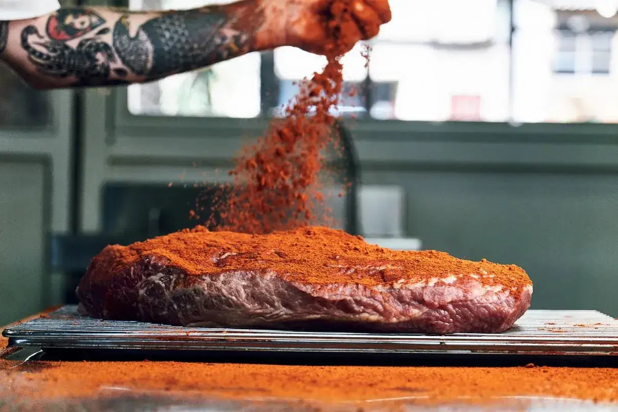 Un trozo de carne cruda, que será pastrami, se cubre con una generosa capa de especias rojas.