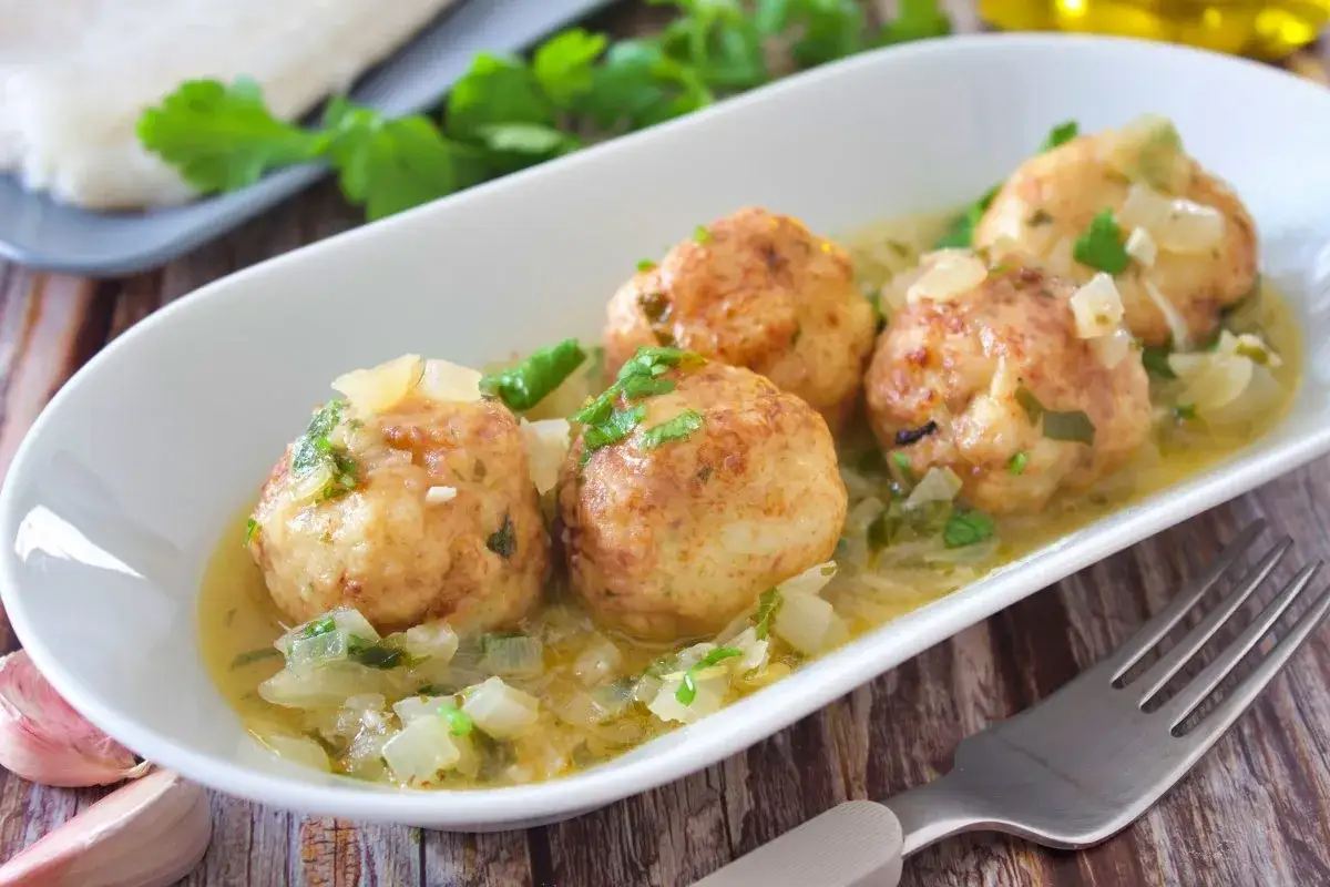 Cuatro albóndigas de merluza caseras en una salsa cremosa con cebolla y perejil, servidas en un plato blanco.