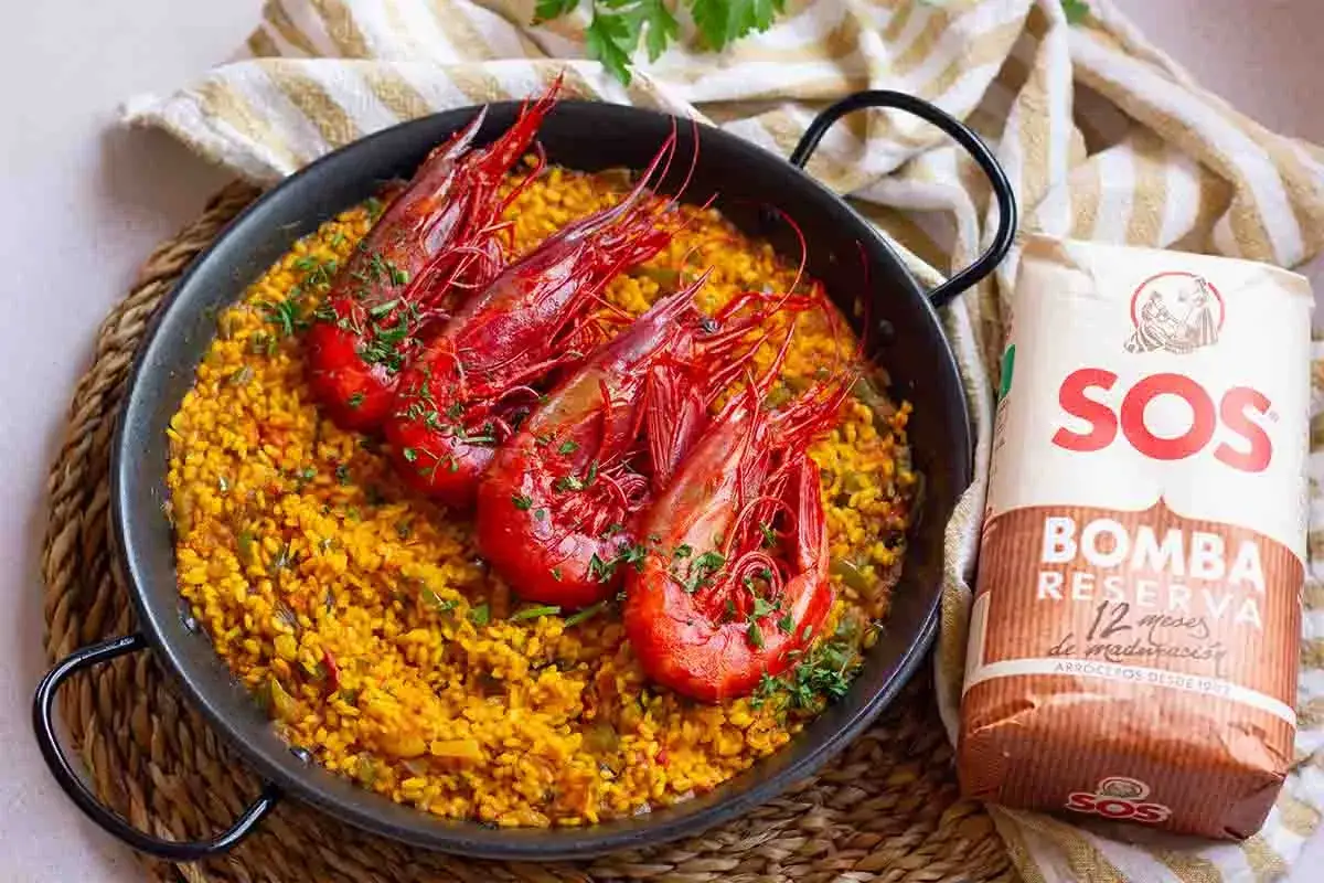 Arroz con carabineros, un plato de paella con gambas rojas y arroz amarillo, junto a un saco de arroz SOS Bomba.