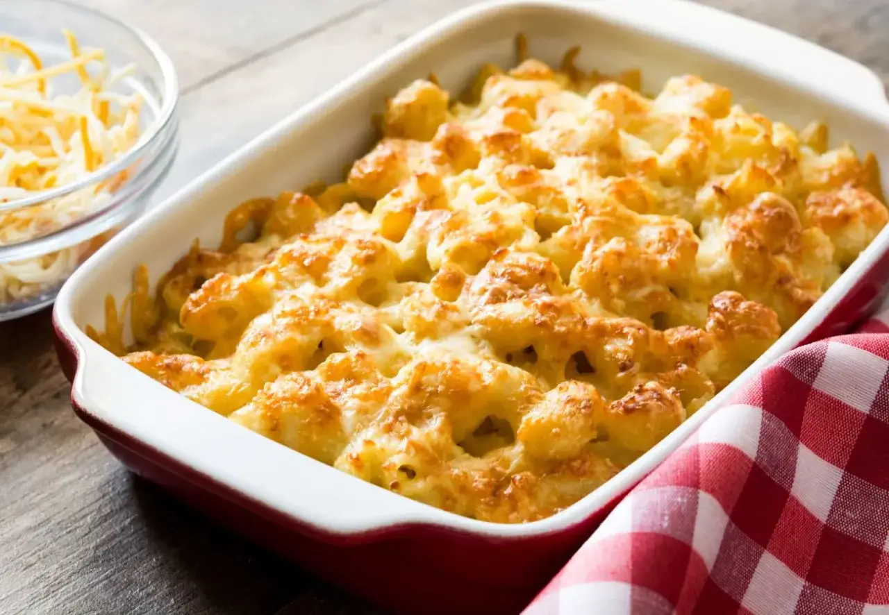 Macarrones con queso reci&eacute;n horneados, dorados y burbujeantes en una fuente roja. Un taz&oacute;n de queso rallado al lado.