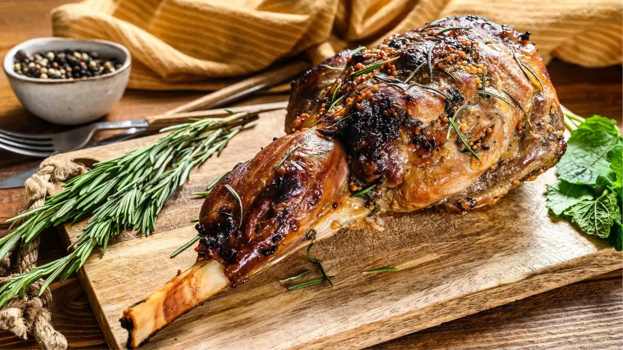 Jugosa pierna de cordero al horno, dorada y aromática, con romero y granos de pimienta. Lista para servir en tabla de madera.