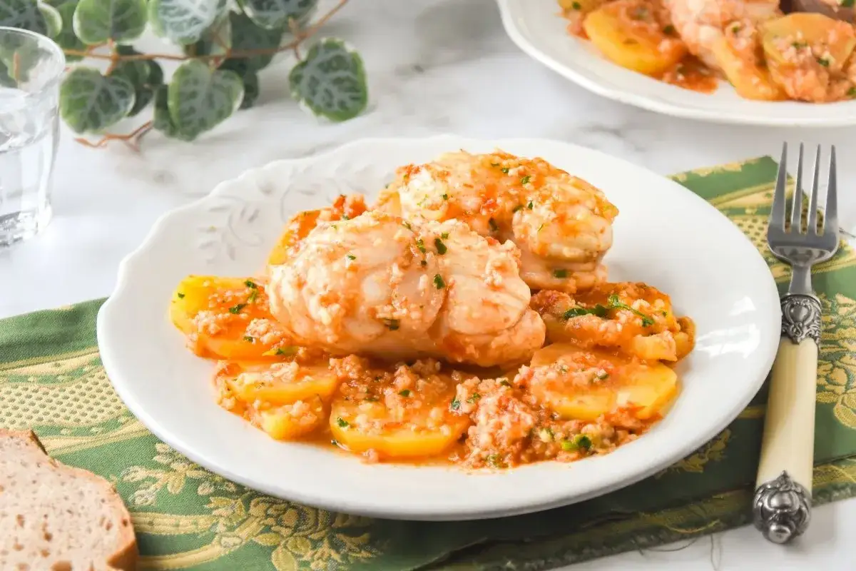 Rape con patatas en salsa de tomate y ajo, servido en plato blanco con pan y tenedor.