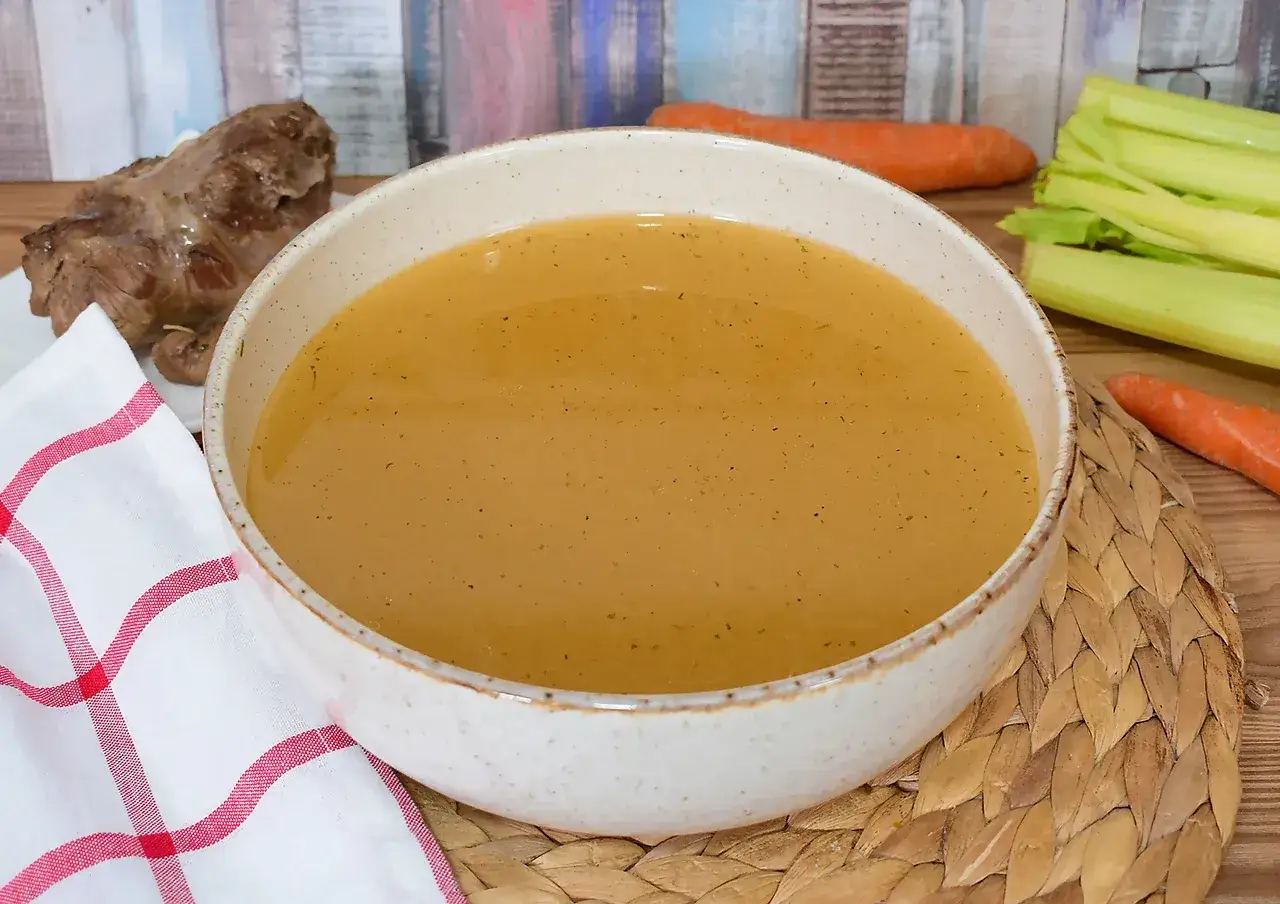 Un tazón de caldo de carne humeante, con un trozo de carne asada y verduras frescas al lado.