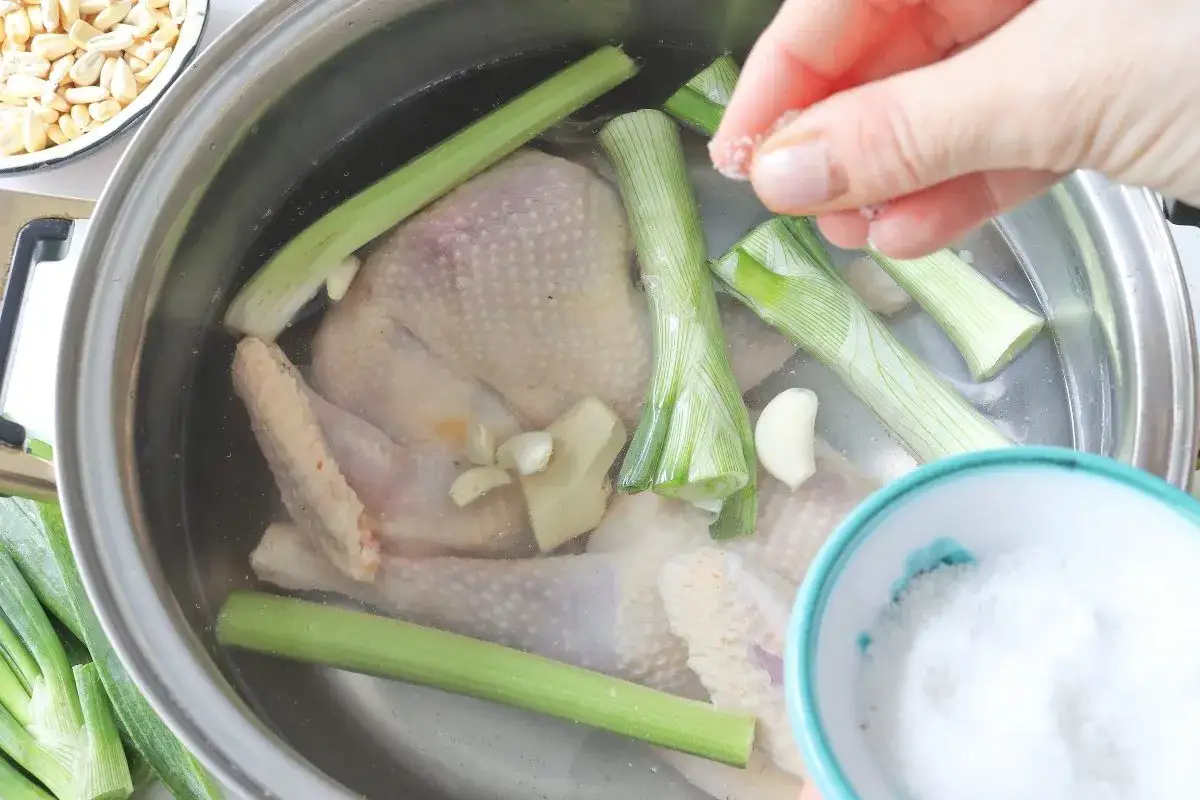 Se añade sal a un pollo entero en una olla con cebolleta, apio y ajo, preparando un delicioso caldo de gallina.