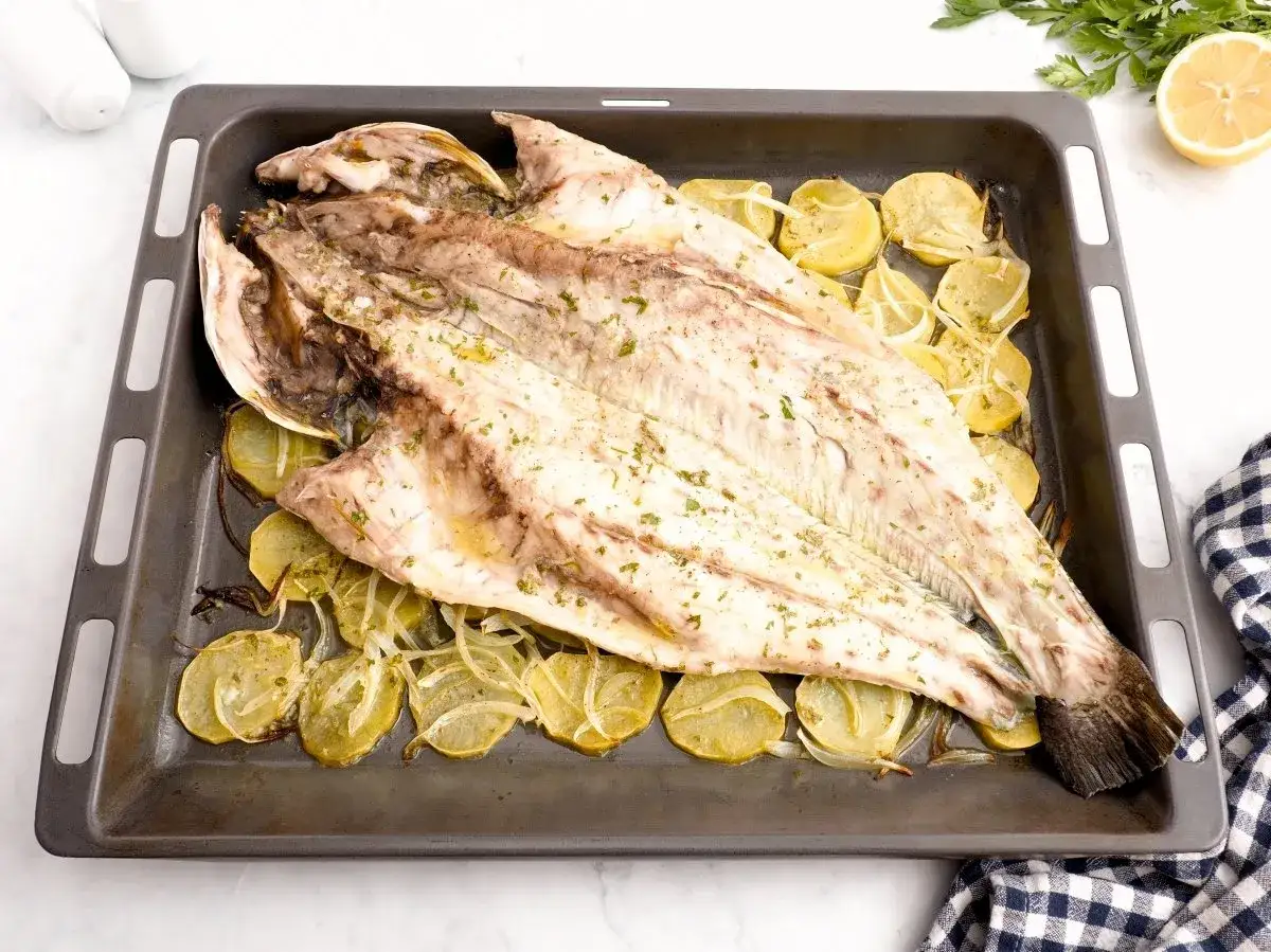 Corvina al horno, entera y dorada, reposa sobre patatas y cebolla en una bandeja. Hierbas frescas espolvoreadas realzan su aroma.