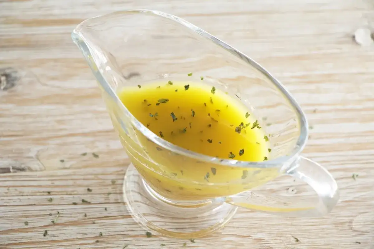 Vinagreta de mostaza dorada con hierbas verdes picadas, servida en una salsera de cristal sobre una mesa de madera.