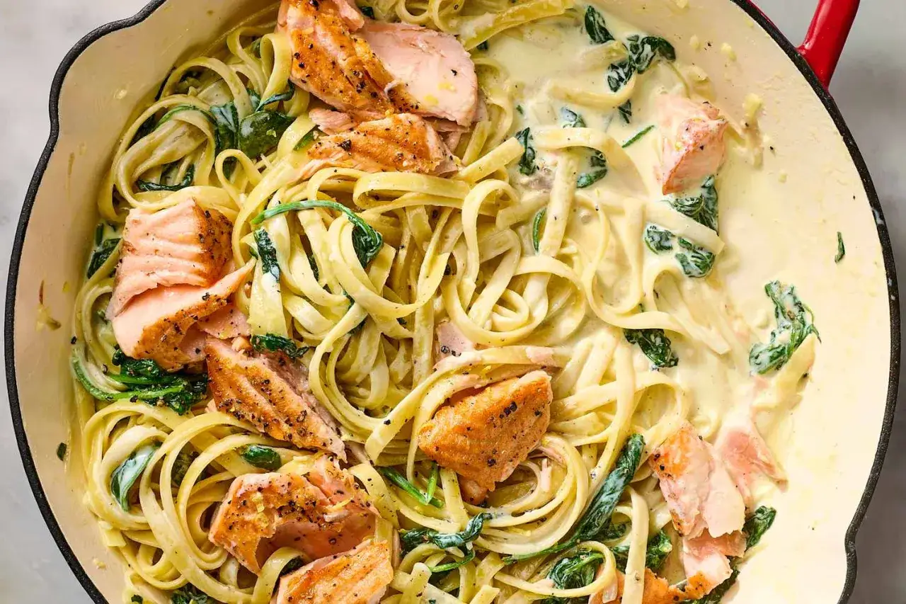 Pasta cremosa con trozos de salmón a la parrilla y espinacas, servida en una sartén.