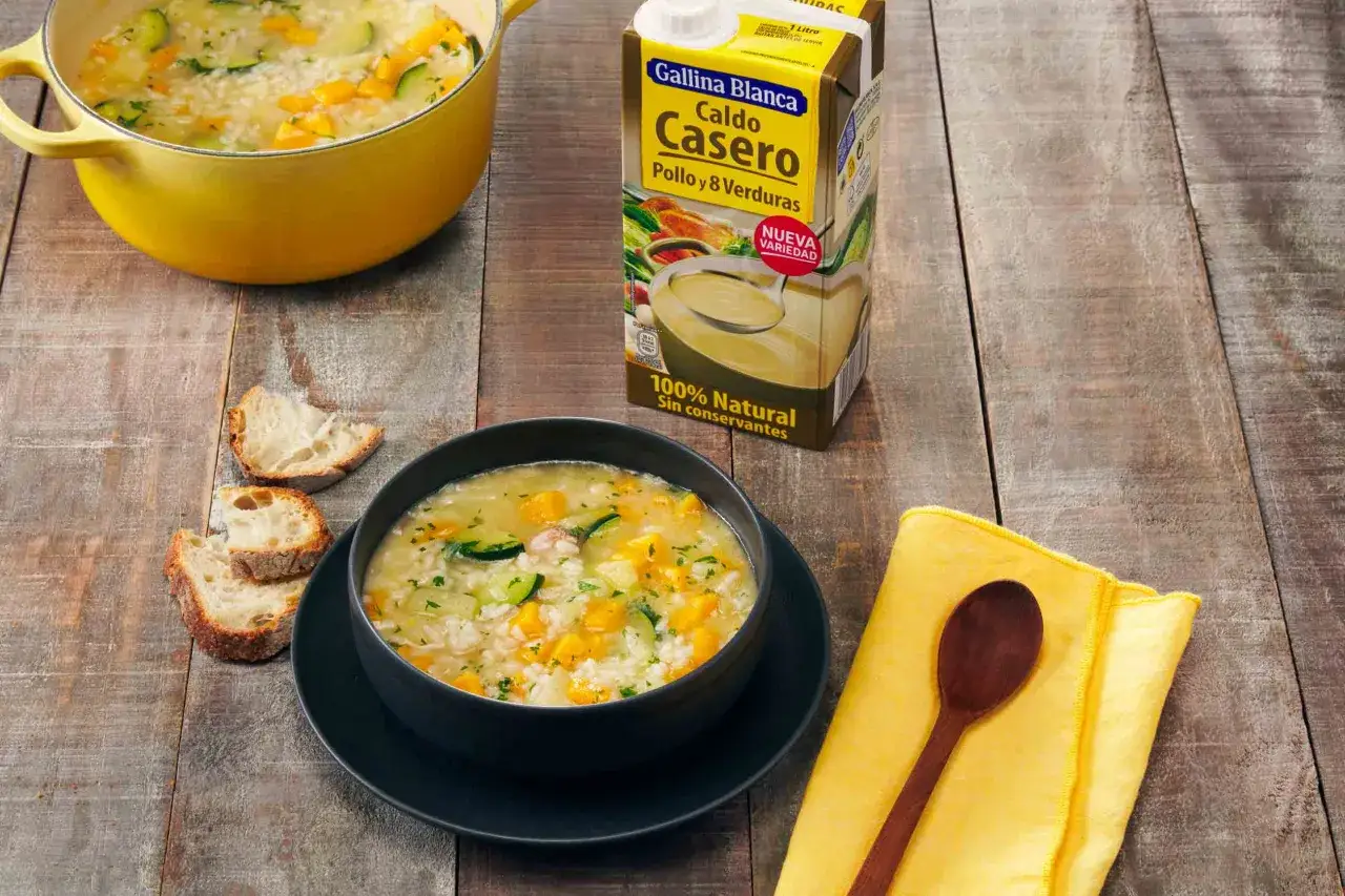 Un plato humeante de sopa de arroz con calabacín y zanahoria, acompañado de pan rústico y un cartón de caldo Gallina Blanca.