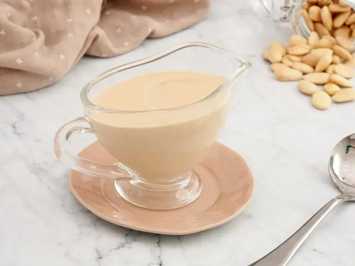 Salsa de almendras cremosa en un recipiente de vidrio, con almendras enteras esparcidas cerca.